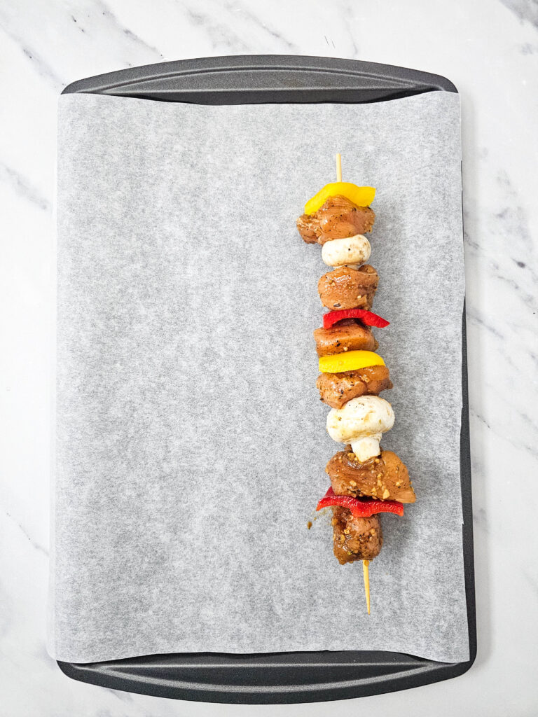 Marinated chicken, bell peppers, and mushrooms threaded onto a skewer on a parchment-lined baking sheet.
