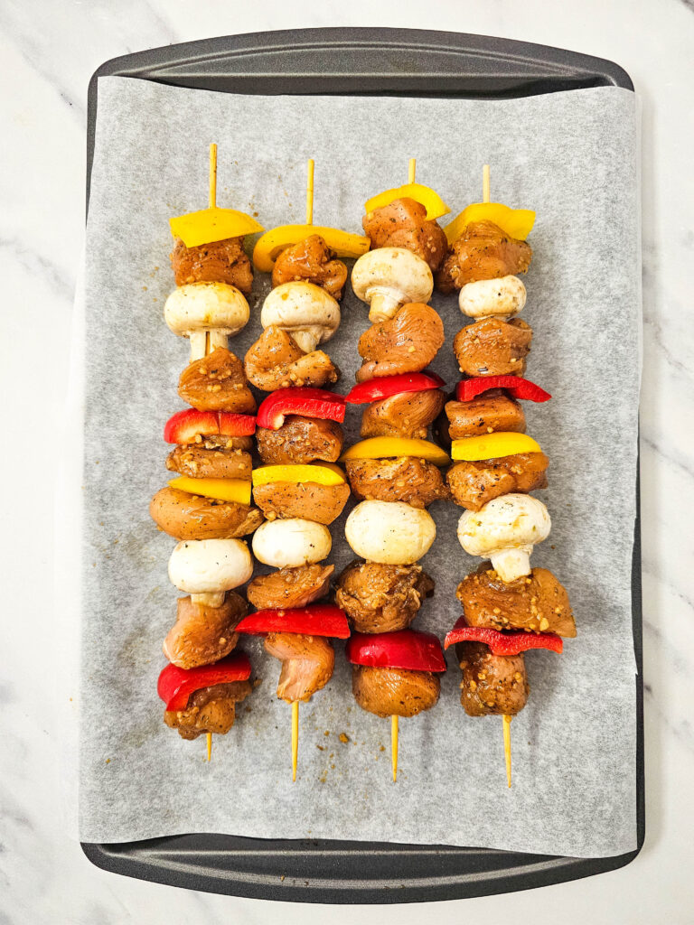 Multiple chicken and vegetable skewers arranged on a parchment-lined baking sheet before cooking.
