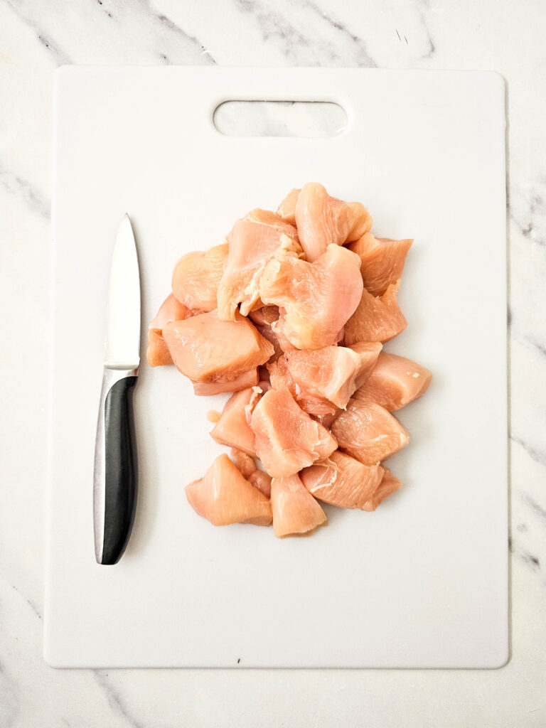 Raw chicken breast cut into bite-sized pieces on a cutting board with a knife.