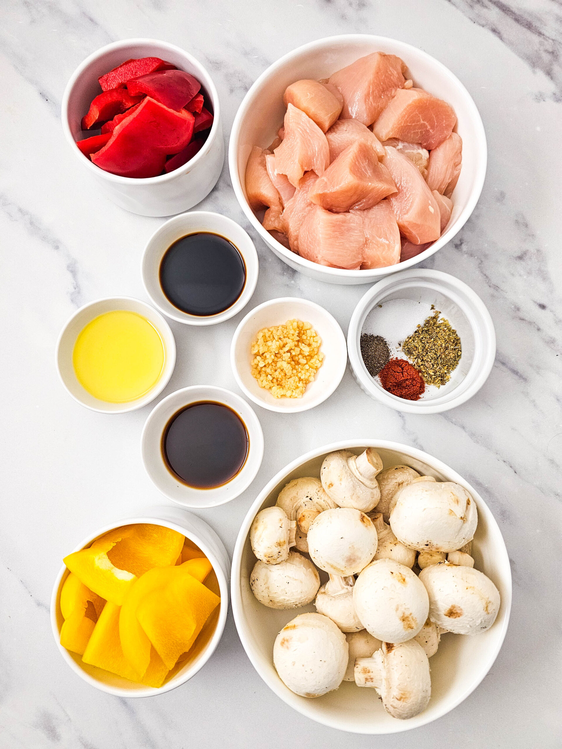 Bowls of raw chicken, chopped bell peppers, mushrooms, oil, sauces, garlic, and spices laid out for prep.